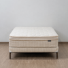 A quilted mattress on a matching upholstered bed frame with dark metal legs, set against a plain light gray wall and wood-look floor.