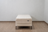 A quilted mattress on a matching upholstered bed frame with dark metal legs, set against a plain light gray wall and wood-look floor.