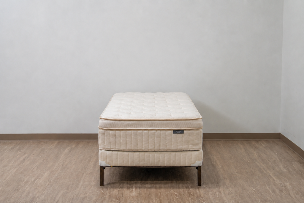 A quilted mattress on a matching upholstered bed frame with dark metal legs, set against a plain light gray wall and wood-look floor.