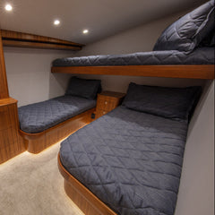 A compact yacht or boat cabin interior with two single beds under a loft bed, upholstered in dark gray quilted bedding, surrounded by warm wood cabinetry and a light beige carpet.